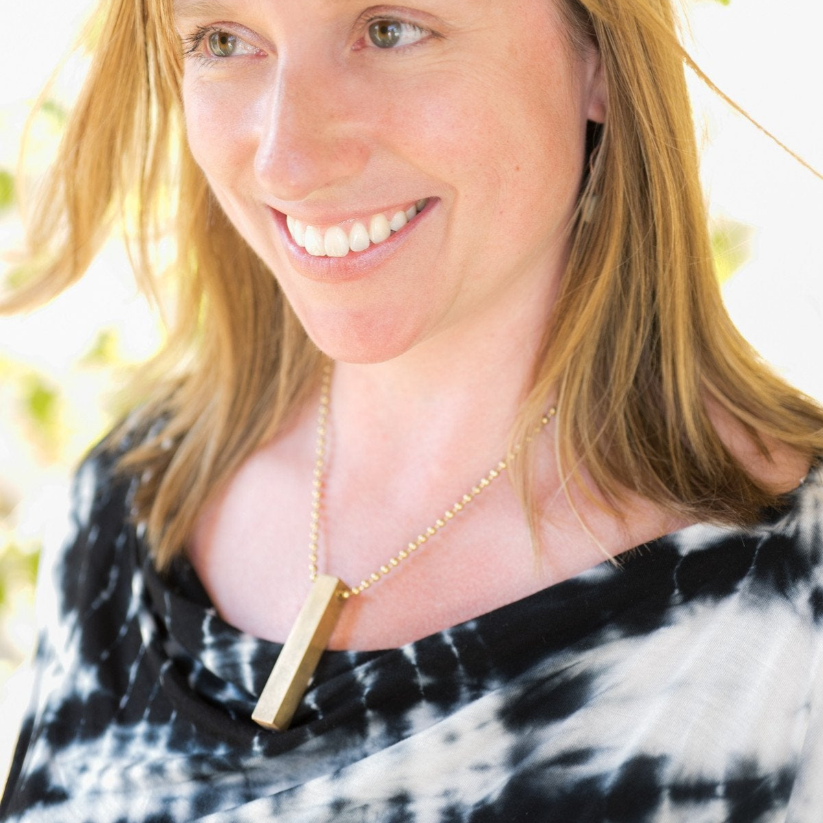 Close-up of a woman's neck and collarbone, wearing the square brass travel dice as a necklace using a longer section of brass beaded chain.  The woman is smiling.